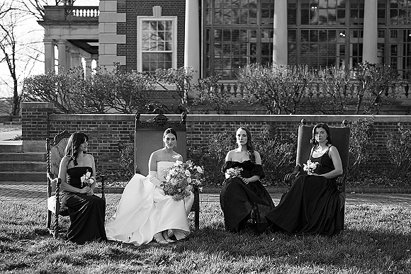 Bridal party portrait of bride with bridesmaids seated on vintage chairs, holding bouquets before a brick building with large windows