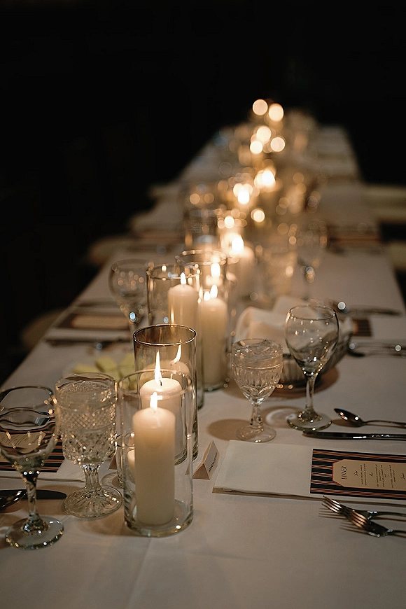 Reception tablescape with a candlelit wedding table set on a long banquet table, featuring white taper candles and crystal stemware in a dark room