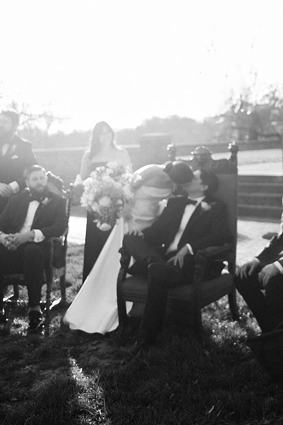 Ceremony kiss at an outdoor wedding ceremony as bride in strapless gown holds a white-green bouquet and groom in tux leans in by chairs on sunlit lawn