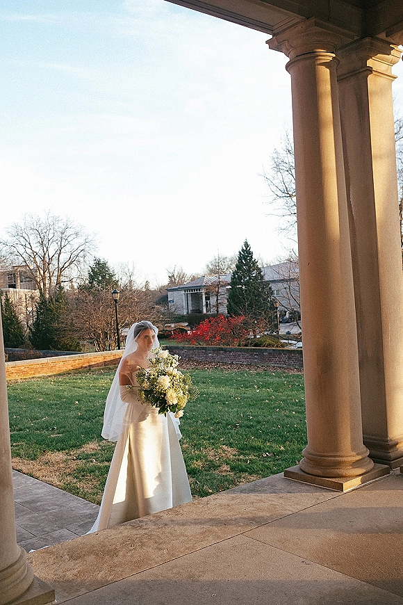 Bridal portrait of a bride holding bouquet in a strapless wedding dress and veil, standing by stone columns on a porch with lawn beyond