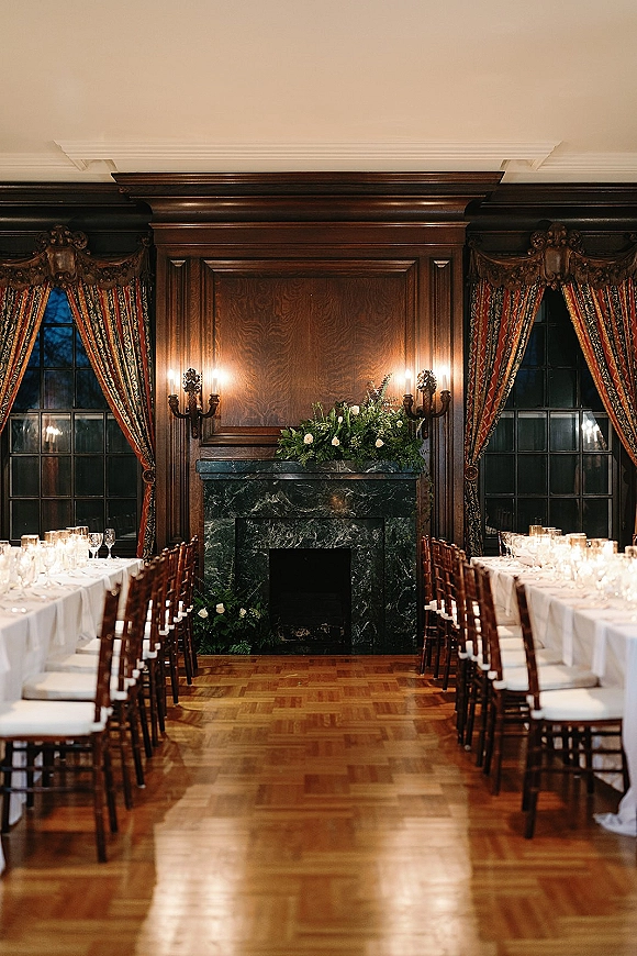 Reception tablescape on a long banquet table with white linens, votive candles, glassware, and mantel greenery by a marble fireplace in a wood-paneled room