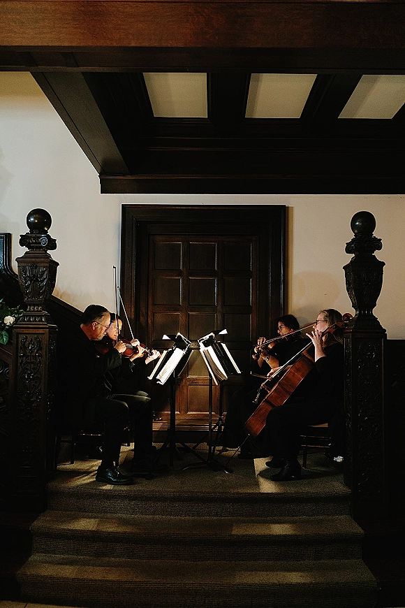 Wedding string quartet in formal black attire playing with sheet music on stands beside a carved staircase in an indoor hallway