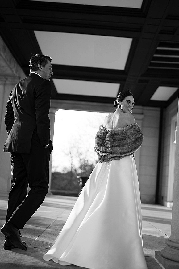 Couple portrait in a black and white wedding portrait style, bride glancing over her shoulder in a fur shawl beside groom in tux on a terrace