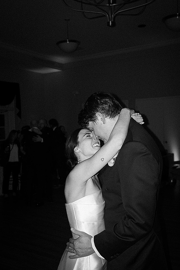 First dance as bride in a strapless wedding dress hugs her groom in a suit on a reception dance floor under a chandelier, guests behind