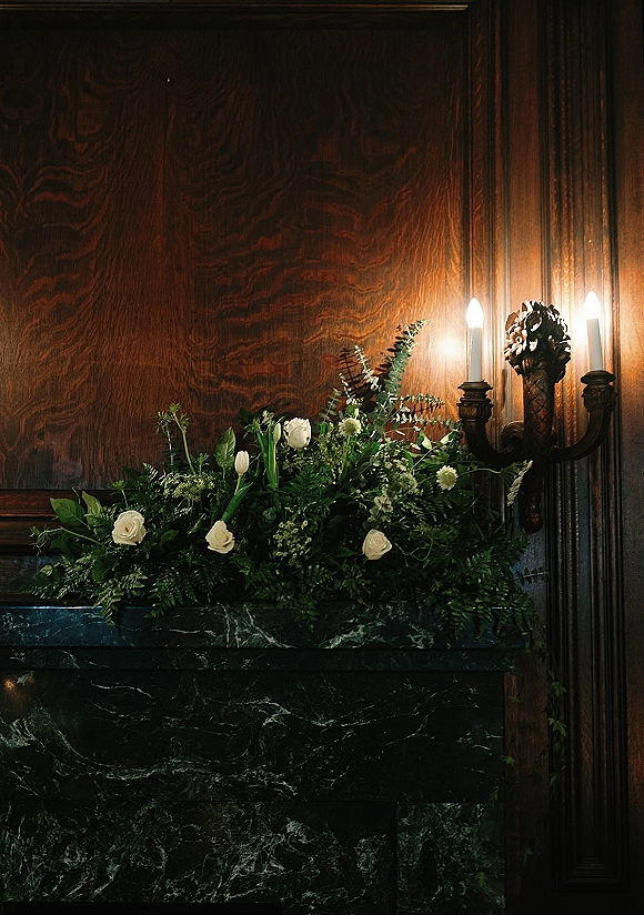 Mantel floral arrangement of wedding mantel flowers with white roses, tulips, and fern greenery garland on a marble fireplace against wood paneling