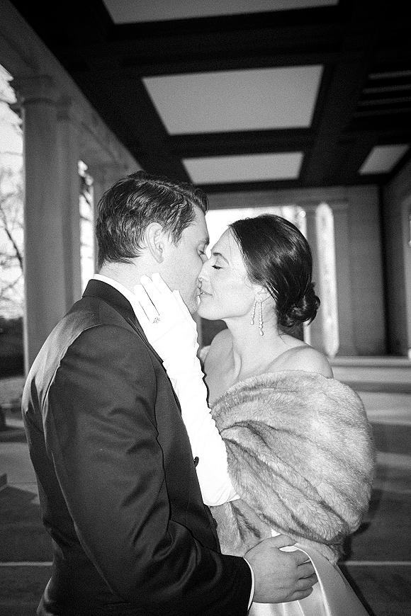 Wedding kiss in a black and white wedding photo, bride in long gloves and fur stole embraces groom in tuxedo under columned walkway