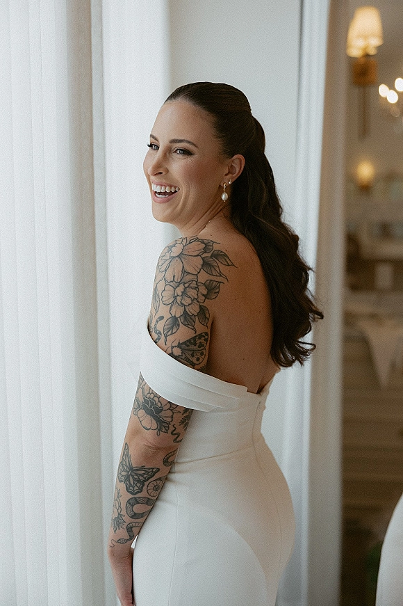 Bridal portrait of a laughing bride in a strapless off-the-shoulder gown, pearl drop earrings, and arm tattoos by window light indoors