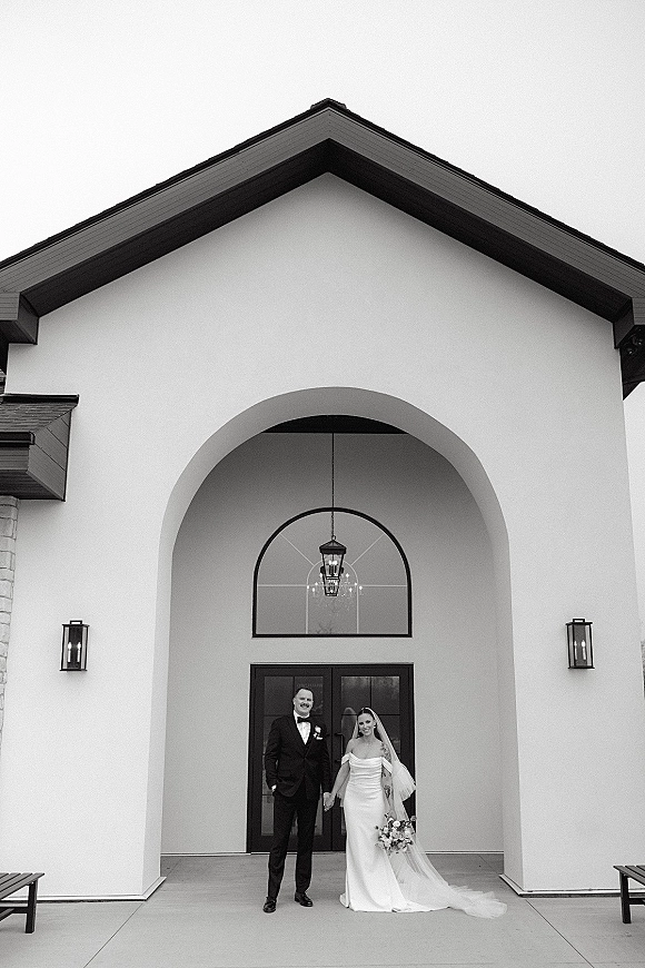 Couple portrait of bride and groom holding hands, her veil and bouquet visible, standing at a chapel entrance beneath an arched doorway