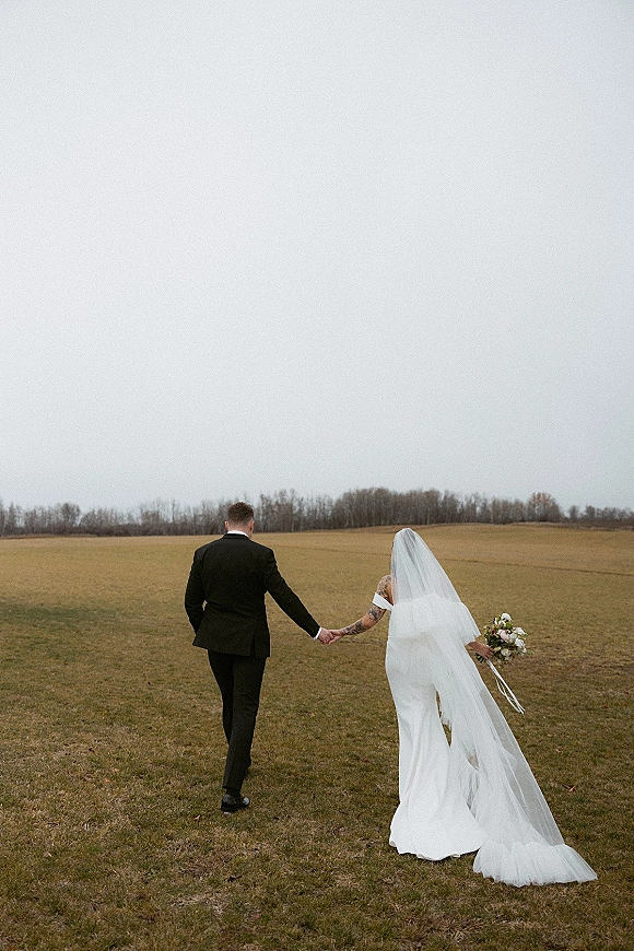 Couple portrait of newlyweds holding hands walking away, bride’s long veil and bouquet trailing in an overcast open field