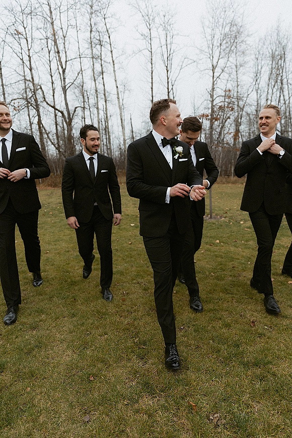 Groomsmen portraits of a wedding party walking and laughing in black tuxedos with bow ties and boutonnieres on a lawn under gray sky