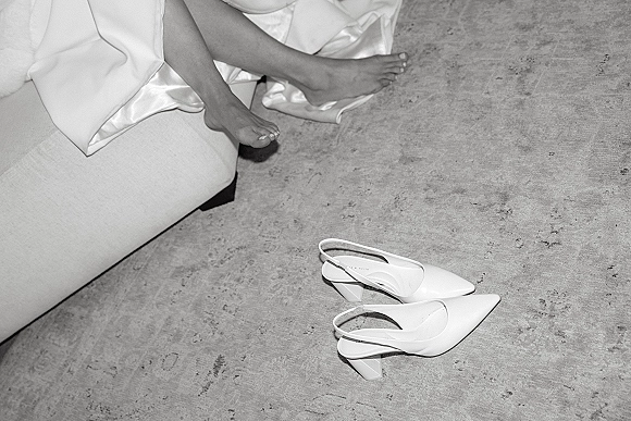 Bridal shoes, white wedding heels in a slingback block-heel style beside a satin bridal robe on a concrete floor near a sofa edge