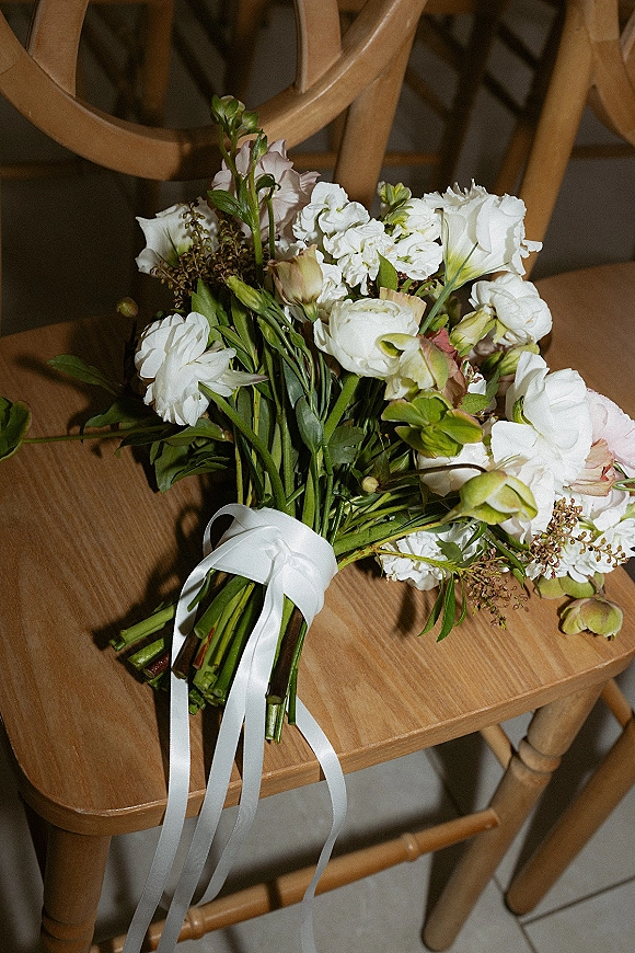 Bridal bouquet of white flowers and greenery with a white ribbon accent resting on a wood chair over a tile floor
