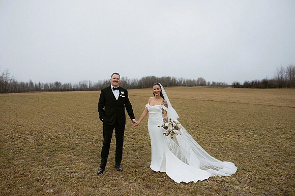 Couple portrait of bride and groom holding hands, her cathedral veil and bouquet flowing beside his tuxedo in an overcast field