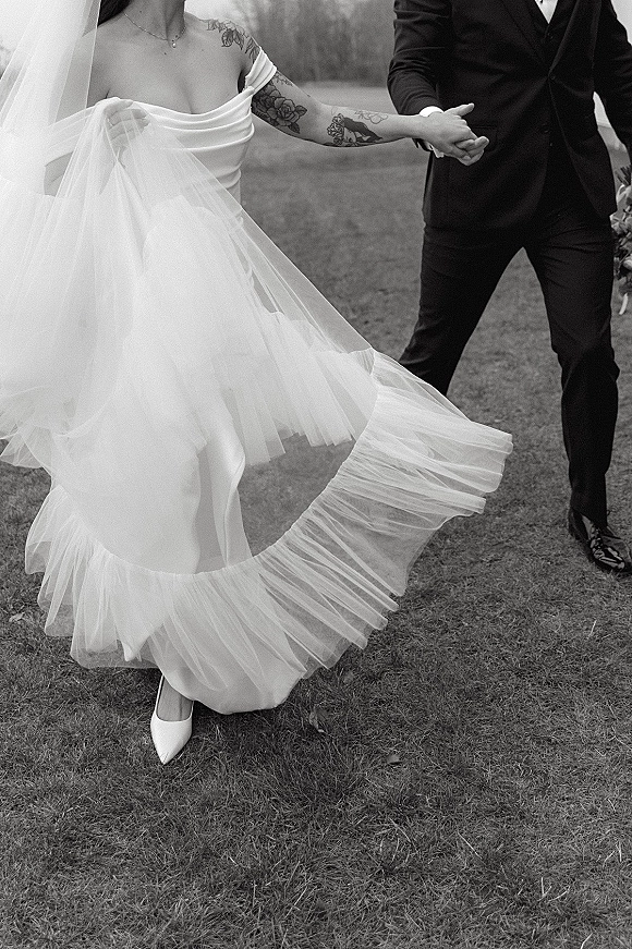 Couple portrait, black and white wedding portrait of bride and groom holding hands as her tulle skirt and veil swirl on a lawn with trees