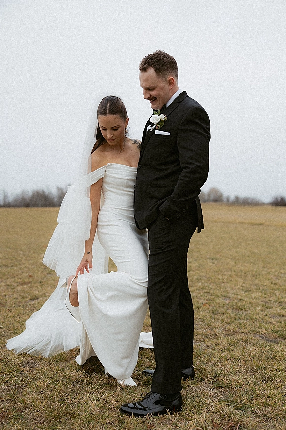 Couple portrait of bride lifting her off-the-shoulder wedding dress to show bridal shoes beside groom in black tuxedo in an open field