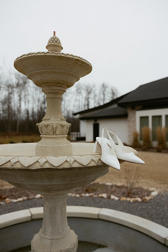 Wedding shoes in white slingback heels resting on a stone fountain in an outdoor courtyard with bare trees under overcast sky