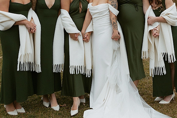 Bridesmaid group photo of bride with bridesmaids holding hands on a grass lawn, in green dresses and white shawls with heels