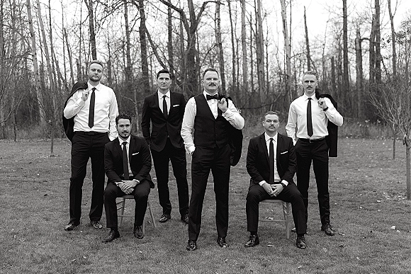 Groomsmen portrait of the groom with groomsmen in suits and vests, some seated on chairs, on a grass lawn with leafless trees behind