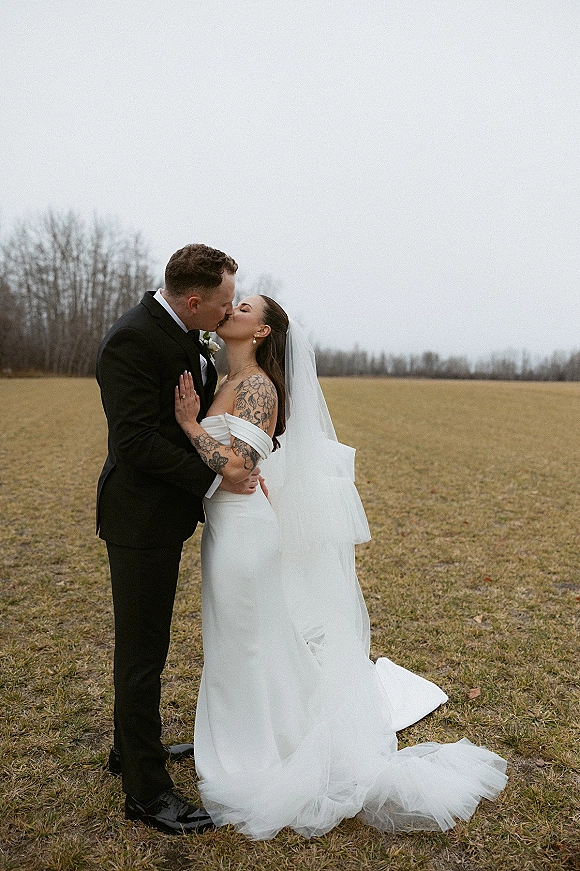 Wedding kiss portrait of bride and groom kissing, her long veil flowing over a strapless dress in an open field under overcast sky