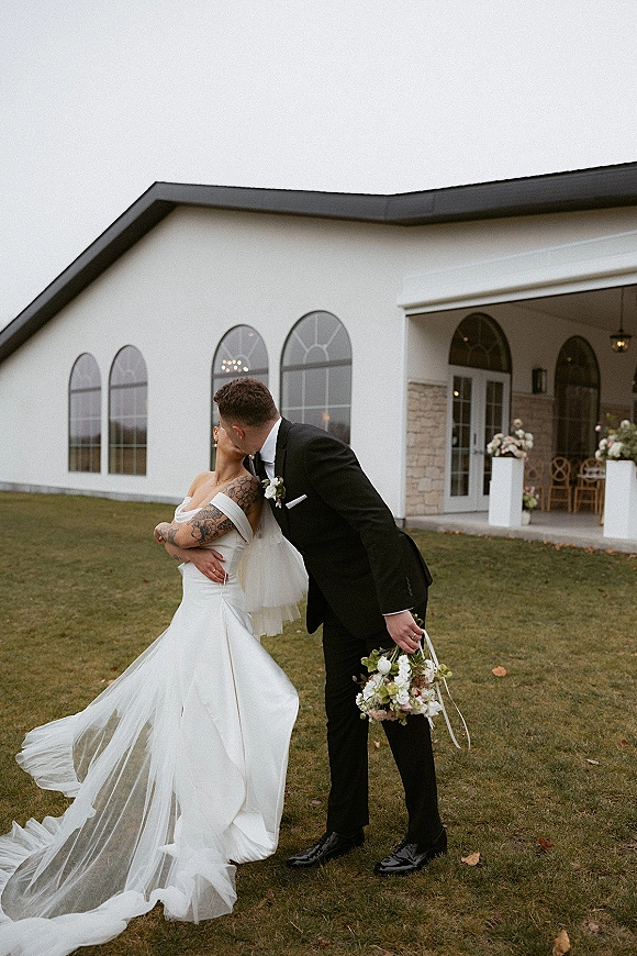 Wedding kiss portrait of bride and groom kissing, her off-shoulder gown and tulle veil flowing beside a white chapel exterior with arched windows