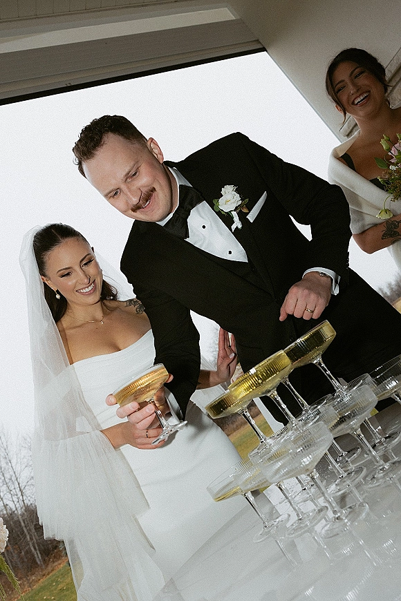 Champagne tower toast as bride in veil and groom in tux pour into gold rim coupe glasses on a table under a patio overhang outdoors