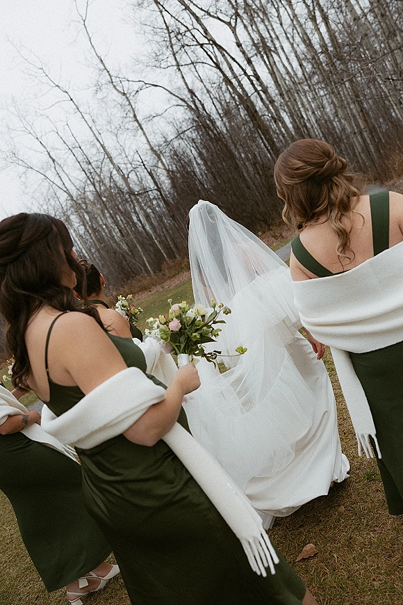 Bride with bridesmaids walking away across a grassy lawn, cathedral veil flowing, dark green dresses with white shawls and bouquets under overcast sky