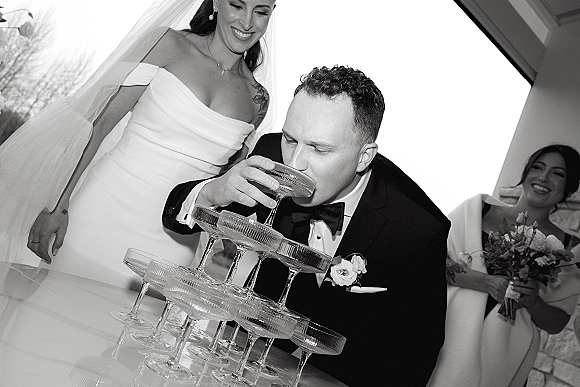 Champagne tower toast as groom sips from a coupe beside the veiled bride in a strapless dress, bright window-lit indoor reception doorway behind