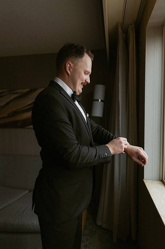 Groom getting ready, groom putting on watch in a black tuxedo with bow tie, standing by window light in a hotel room