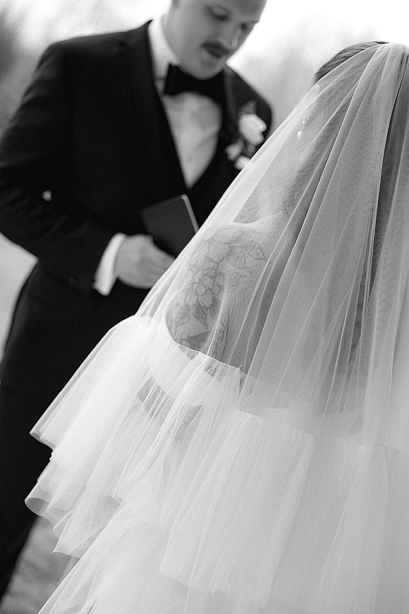 Wedding vows as groom reading vows from a vow book beside bride in bridal veil, tuxedo and boutonniere against open sky backdrop