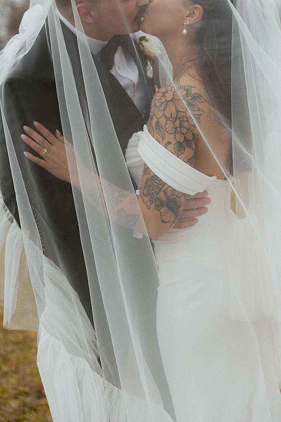 Wedding kiss portrait of bride and groom kissing beneath a sheer veil, her hand on his tuxedo chest in an outdoor field sky