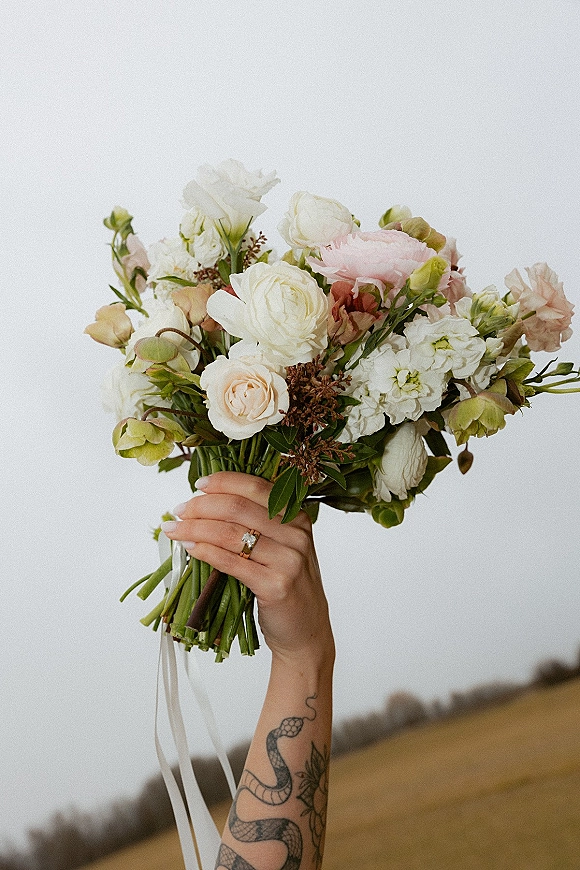 Bridal bouquet with garden style blooms, white roses, blush flowers, and greenery tied with ribbon, held up against a field and sky