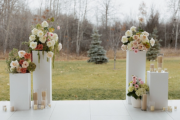 Ceremony aisle decor with white pedestal plinths topped in rose and hydrangea florals, lined with glass cylinder candles on a lawn