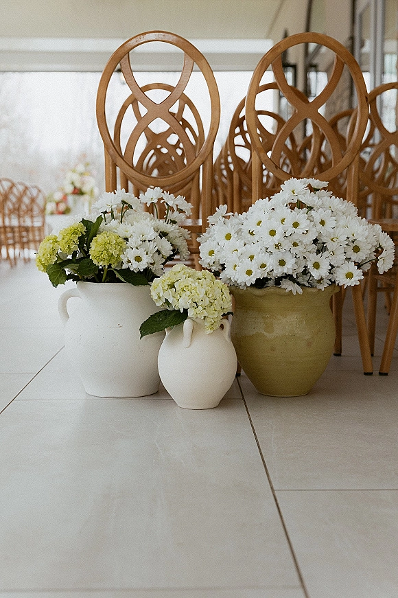 Ceremony aisle decor with aisle floral arrangements of white daisies and green hydrangea in ceramic floor vases beside wooden chairs indoors