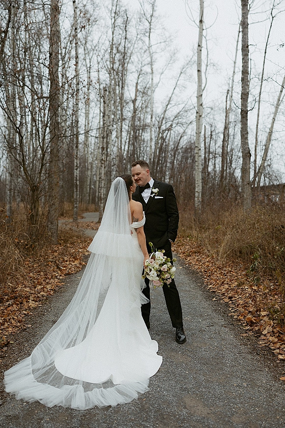 Couple portrait of bride and groom kiss on a forest path, her cathedral veil trailing behind as they hold a pastel bouquet amid fallen leaves
