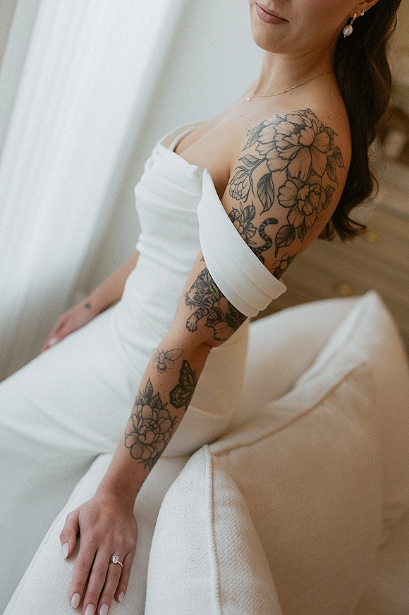 Bridal portrait of a tattooed bride in a strapless dress, showing floral arm tattoos, pearl earrings and ring by sheer curtains indoors