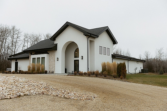 Wedding venue exterior with a stone facade and arched entryway, wall lanterns, and gravel driveway bordered by river rock landscaping under overcast sky