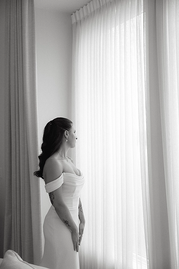 Bridal portrait of a bride by window in an off shoulder gown, side profile with jewelry and tattoos framed by sheer curtains in a bedroom