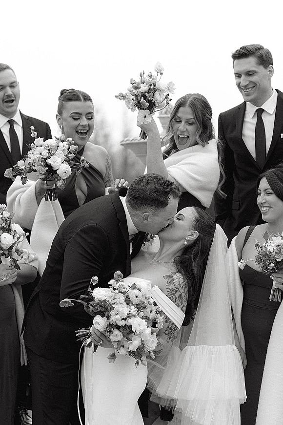 Wedding kiss portrait of bride and groom kissing as wedding party cheers, bride holding white bouquet and veil outdoors under overcast sky