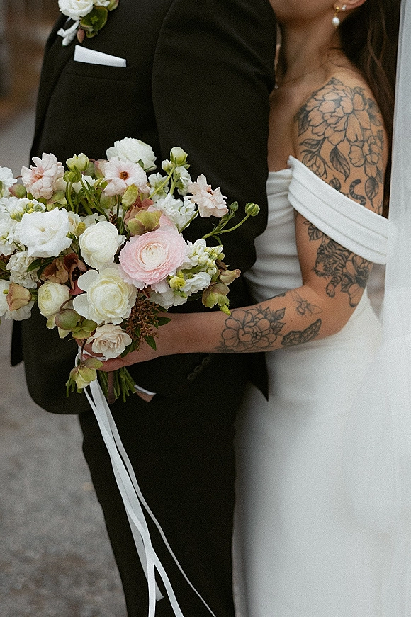 Wedding couple portrait of bride and groom embrace, holding a bridal bouquet with long ribbon against a neutral outdoor wall on pavement