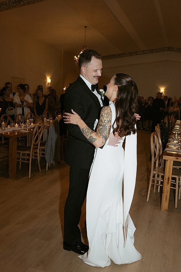 First dance as bride in a white wedding dress and groom in a tuxedo sway on a wood floor under chandelier light, guests watching