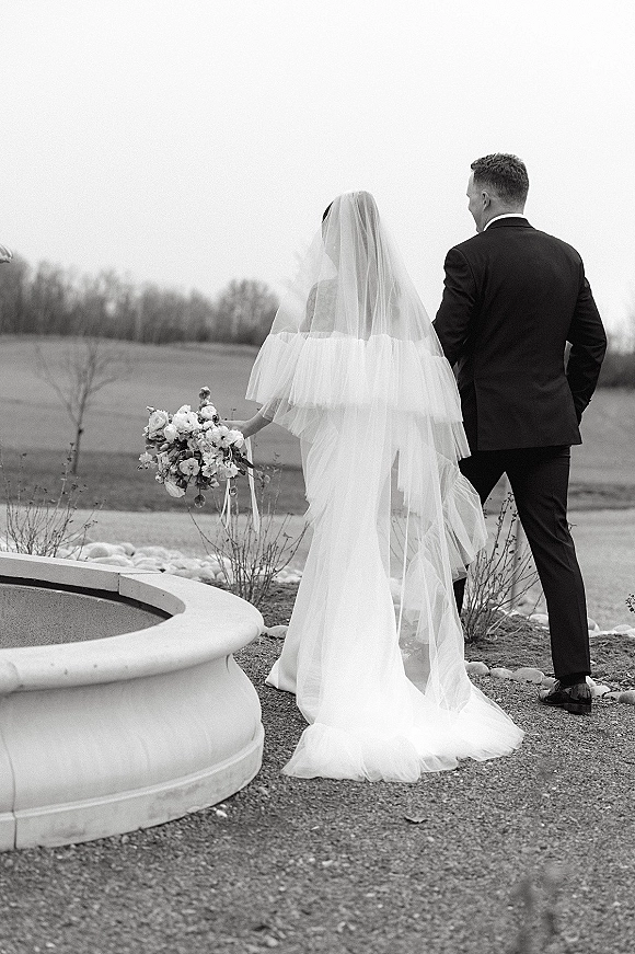 Couple portrait in black and white wedding portrait style, back view bride and groom walking down a gravel path past a fountain, veil trailing