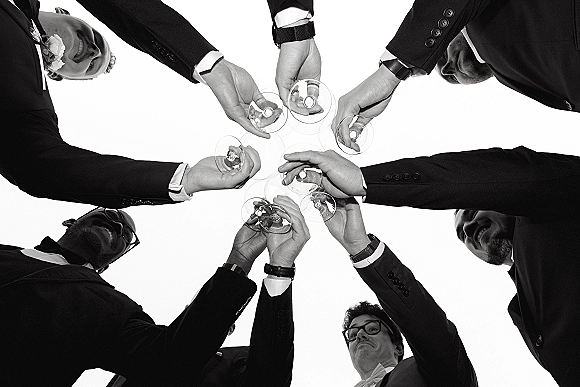 Groomsmen toast with champagne flutes raised, tuxedos and boutonnieres visible as they huddle in a circle against a bright sky