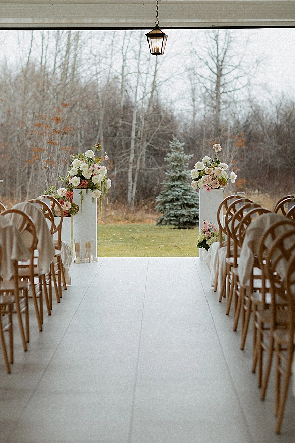 Ceremony aisle decor with a wedding aisle runner, wooden chairs draped in fabric, floral pedestal stands, and glass cylinder candles on a covered porch