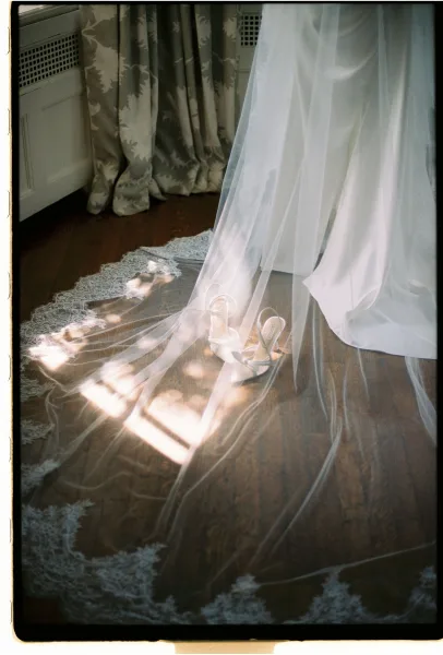 Wedding veil with cathedral veil lace trim spread on a wood floor beside white bridal heels, softly lit by window light and patterned curtains