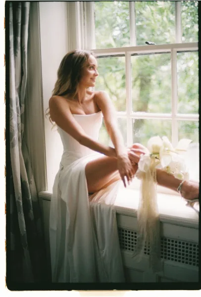 Bridal portrait of a bride by window, seated on a window seat in soft natural light, holding a white orchid bouquet with long ribbon accent