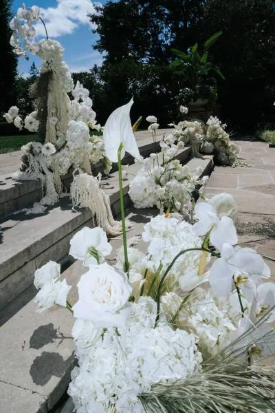 Ceremony floral decor of lush white orchids and hydrangeas in urns and grounded clusters framing stone steps on a garden patio