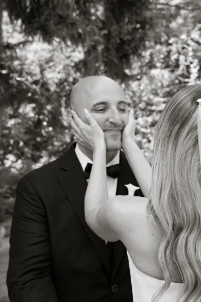 Couple portrait of bride holding groom’s face as he smiles, her veil and strapless dress beside his tuxedo in lush greenery