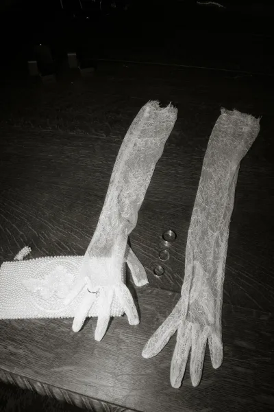 Wedding rings in a wedding ring flat lay beside lace bridal gloves and a pearl clutch purse on a dark wood floor