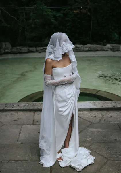 Bridal portrait of a bride in a strapless wedding dress with lace veil over face, standing by a stone fountain in a garden courtyard
