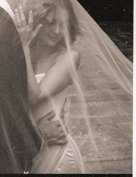 Bridal portrait of a bride with veil draped over her face, strapless gown and wedding ring visible against a dark, moody backdrop